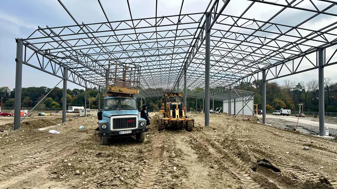 Steel frame of a future warehouse over a compacted ground pad, with a grader working nearby